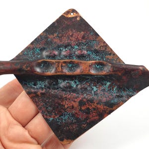 Puede incluir: Un colgante de pared de cobre con forma geométrica y superficie texturizada. El cobre tiene un acabado de pátina con tonos verdes, marrones y rojos.