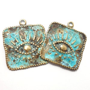 Peut inclure: Deux breloques carrées en laiton avec un motif d'œil. Les breloques sont de couleur bleu turquoise avec des accents dorés. Le motif de l'œil a une pupille dorée et des cils.
