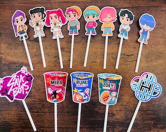 May include: A collection of colorful cake toppers featuring cartoon characters and ramen cup designs. The toppers include various characters with different hairstyles and outfits, along with ramen cup illustrations with text. The toppers are on white sticks.