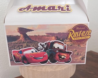 May include: White gable box with a handle, featuring a Cars theme. The box has a colorful image of Lightning McQueen and Mater, with the text "Amari" on top. A "Rusteze" sticker is also visible.