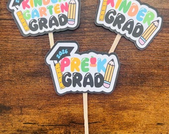 May include: Three graduation cake toppers. Two toppers read "Kindergarten Grad" with a graduation cap, ruler, and pencil. The third topper reads "Pre-K Grad" with a graduation cap, apple, ruler, and pencil. All toppers have the year 2026.
