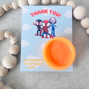 May include: A light blue birthday card with the text "THANK YOU!" and images of Spider-Man characters. An orange plastic container is in front of the card. The card also says "FOR COMING TO MY BIRTHDAY! NAME".