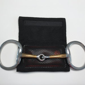 May include: A silver and gold horse bit with a black mesh bag. The bit has two rings and a jointed mouthpiece.