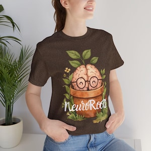May include: A brown t-shirt with a playful design. It features a brain wearing glasses, planted in a terracotta pot, with the text "NeuroRoots". The design includes green leaves and small yellow flowers, creating an educational theme.
