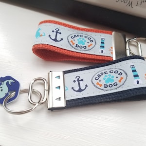 May include: Two keychains with a nautical theme. One has an orange strap, the other is navy blue. Both have a light blue band with anchors, lighthouses, and the text "Cape Cod Dog". A blue key with a whale design is attached.
