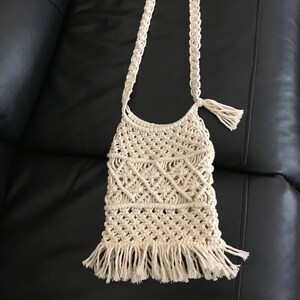 May include: A white crocheted bag with a long strap and fringe detail. The bag is made of cotton cord and has a bohemian style.