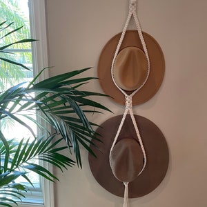May include: A white macrame hat rack with two hats hanging on it. The hats are brown and beige. The rack is hanging on a white wall.