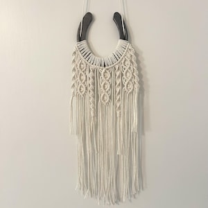 May include: A wall hanging featuring a dark metal horseshoe wrapped in off-white macrame cord. The cord is knotted in intricate patterns, including vertical braids and diamond shapes, ending in long, loose fringe strands hanging downward.