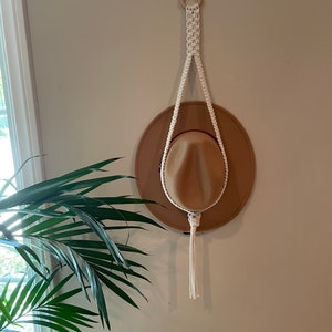 May include: A white macrame hat hanger with a wooden ring at the top and a tassel at the bottom. A brown hat is hanging on the hanger.