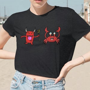 Puede incluir: Una camiseta negra recortada con un diablo rojo de dibujos animados sosteniendo un tridente y un cangrejo rojo de dibujos animados. La camiseta la lleva puesta una persona en una playa.