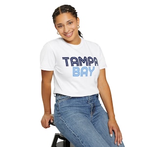 Könnte beinhalten: Ein weißes kurzärmeliges T-Shirt mit Rundhalsausschnitt und dem Schriftzug TAMPA BAY auf der Brust. Das Wort TAMPA ist in Dunkelblau mit einer dreifachen Linienkontur gehalten, das Wort BAY in Hellblau mit einem dreifachen Liniendesign.