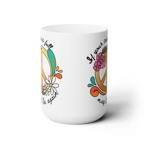May include: White ceramic mug featuring a peace sign design. The peace sign is yellow with colourful flowers and swirls. The text on the mug reads "If your cup is full, may it be again."