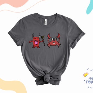 Puede incluir: Una camiseta gris oscuro con un estampado gráfico de un diablo rojo y un cangrejo rojo. El diablo sostiene un tenedor y tiene la lengua afuera. El cangrejo tiene ojos grandes y sonríe. La camiseta está anudada en la parte inferior.