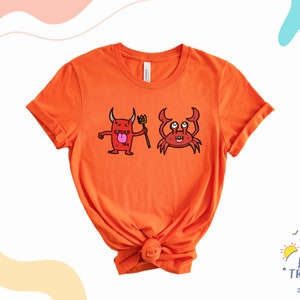May include: Orange t-shirt with a red cartoon devil holding a trident and a red cartoon crab. The shirt is knotted at the bottom.