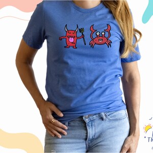 Puede incluir: Una camiseta azul real con un diablo rojo sosteniendo un tenedor y un cangrejo rojo con ojos grandes. La camiseta tiene el texto "Happy Traveler Designs" en la esquina inferior derecha.