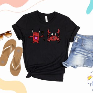 Puede incluir: Camiseta negra con un gráfico de un diablo rojo y un cangrejo. La camiseta se muestra con sandalias marrones, gafas de sol y pantalones cortos de mezclilla.