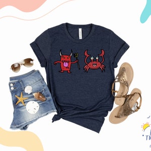 May include: A navy blue t-shirt with a red devil and a red crab graphic. The devil is holding a trident and has a tongue sticking out. The crab has big eyes and is smiling. The t-shirt is on a white background with a pair of sunglasses, denim shorts, and sandals.