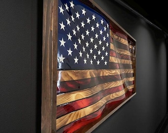 May include: Wooden American flag wall art. The flag features a blue canton with white stars and red and white stripes. The wood has a natural grain and is encased in a dark brown frame. The flag is mounted on a dark gray wall.