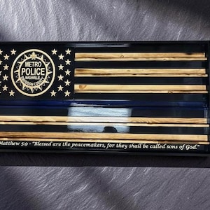 May include: A rectangular wooden wall plaque featuring a stylized American flag design with natural wood stripes and a black canton. The canton displays the Metro Police Nashville Davidson County badge and stars. A blue stripe runs horizontally, and the text Matthew 5:9 - Blessed are the peacemakers, for they shall be called sons of God is printed at the bottom.