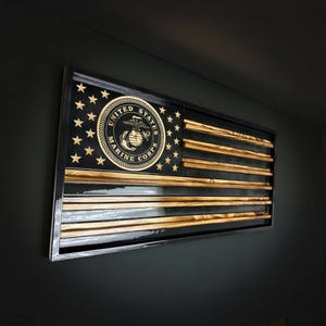 May include: A rectangular wooden wall art piece featuring a stylized United States flag design. The canton is black with gold stars and a central United States Marine Corps emblem. The stripes alternate between natural wood grain and black.