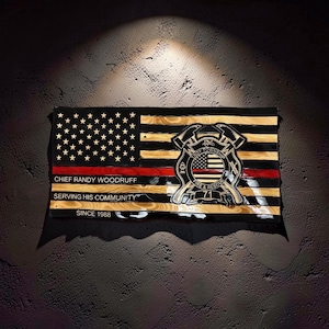 May include: A rectangular wooden wall sign featuring a stylized American flag with black and natural wood stripes and a thin red line. It displays the text CHIEF RANDY WOODRUFF, SERVING HIS COMMUNITY, SINCE 1988, and a Troy Township Fire & Rescue 71 emblem.