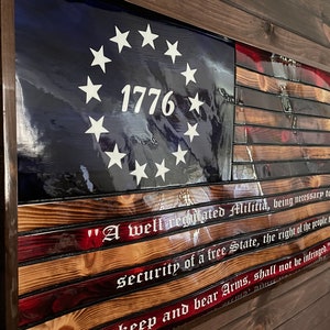 May include: A wooden American flag with 13 red and white stripes and a blue field with 13 white stars in a circle. The year 1776 is printed in white on the blue field. The text "A well regulated Militia, being necessary to the security of a free State, the right of the people to keep and bear Arms, shall not be infringed." is printed in white on the bottom of the flag.