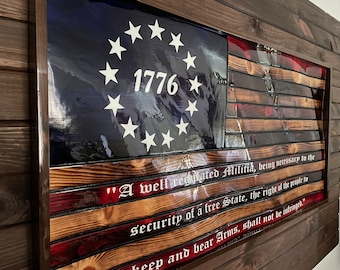 May include: A wooden American flag with 13 red and white stripes and a blue field with 13 white stars in a circle. The year 1776 is printed in white on the blue field. The text "A well regulated Militia, being necessary to the security of a free State, the right of the people to keep and bear Arms, shall not be infringed." is printed in white on the bottom of the flag.