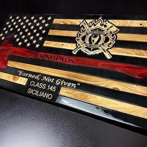 May include: A rectangular wooden wall plaque featuring a stylized American flag with alternating black and natural wood stripes. A red axe is centered across the flag with the text PICKHEADS, a fire academy emblem, and the text Earned, Not Given, CLASS 145, SICILIANO.
