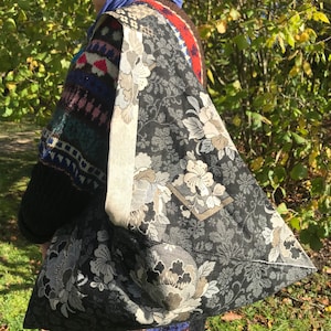 May include: A large, black tote bag with a floral pattern in shades of gray and cream. The bag has a wide strap and is carried over the shoulder. The background shows green foliage.