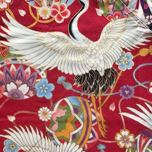 May include: Close-up of a red fabric with a Japanese crane design. The fabric features white cranes with gold accents, surrounded by floral and geometric patterns in blue, purple, and gold. The background is a textured red.