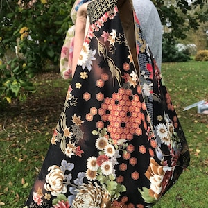 May include: A black tote bag with a floral pattern featuring white, red, and brown flowers. The bag has a geometric design in gold and red. The bag is held by a person outdoors.