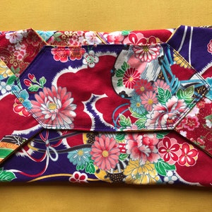 May include: A folded clutch with a floral pattern in red, purple, and white. The clutch features a geometric design with a flap closure. The fabric is adorned with various flowers and leaves.