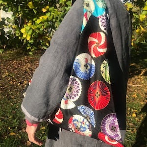 以下が含まれることがあります： カラフルな日本の傘模様の黒いトートバッグ。バッグは肩にかけられ、グレーのジャケットとカラフルなスカーフが見えます。傘は赤、青、紫、白です。