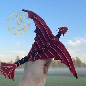 Puede incluir: Un juguete de dragón rojo y dorado hecho a mano, con alas extendidas. El dragón tiene detalles dorados y una columna vertebral negra. El dragón se sostiene contra un cielo azul y un logotipo con una aguja e hilo.