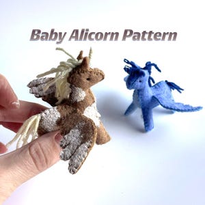 Peut inclure: Deux petites figurines alicorne en feutrine avec des ailes et des crinières en laine. L'une est marron avec des taches blanches et de la laine crème, l'autre est bleu uni. Le texte Baby Alicorn Pattern est affiché au-dessus.