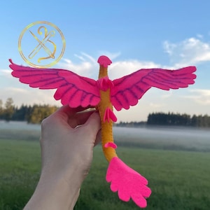 Könnte beinhalten: Ein handgefertigtes Drachenspielzeug aus Filz mit leuchtend rosa Flügeln und Schwanz und einem gelben Körper. Der Drache wird vor dem Hintergrund eines grünen Feldes und eines blauen Himmels gehalten. Die Flügel sind ausgebreitet und der Schwanz ist gebogen.