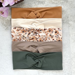 Könnte beinhalten: Set aus fünf geknoten Stirnbändern in verschiedenen Farben und Mustern. Die Stirnbänder sind aus einem weichen, dehnbaren Stoff gefertigt und perfekt, um die Haare aus dem Gesicht zu halten. Das Set enthält ein braunes Stirnband, ein beiges Stirnband, ein Stirnband mit Blumenmuster, ein braunes Stirnband und ein grünes Stirnband.