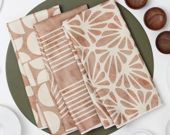 May include: Set of four kitchen towels with different geometric patterns in shades of brown and beige. The towels are folded and arranged on a green surface.