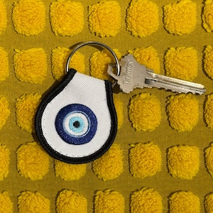 May include: A white and black Evil Eye keychain with a silver key attached. The keychain has a blue eye design in the center. The background is a yellow textured fabric.