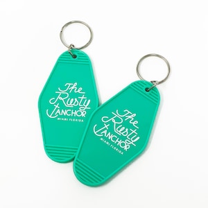 May include: Two teal keychains with silver key rings. The keychains have a retro motel style shape and feature the white text "The Rusty Anchor Miami Florida". The keychains are on a white background.