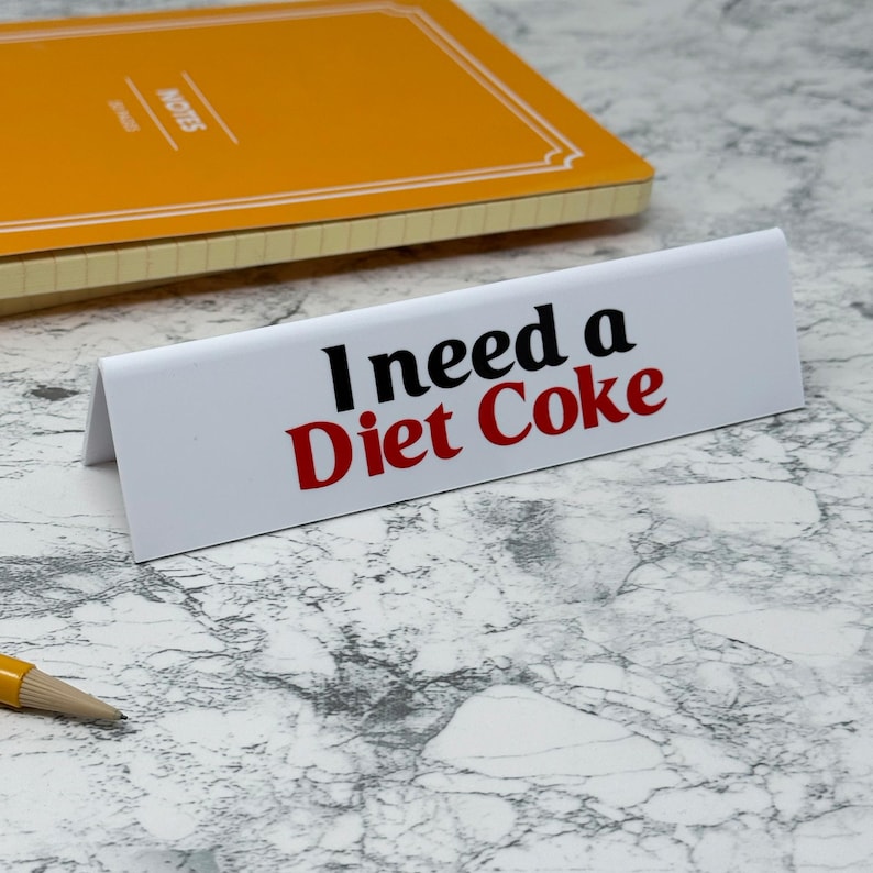 May include: A white desk sign with the text "I need a Diet Coke" in black and red. An orange notebook is in the background, along with a pencil. The sign is angled, and the background is a marble pattern.