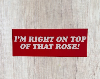 Op de afbeelding: Een rechthoekig rood bord met de witte tekst "I'M RIGHT ON TOP OF THAT ROSE!". De tekst in hoofdletters en vetgedrukt komt goed uit tegen een lichte houtnerf achtergrond.