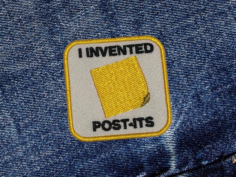 May include: A yellow and white embroidered patch with the text "I invented Post-its" and a yellow sticky note illustration.