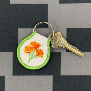May include: A key ring with a white, teardrop-shaped patch featuring an embroidered design of orange poppies with green stems and leaves. The patch is outlined with a green border. A silver key ring holds the patch and a gold-coloured key.