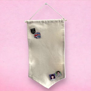 May include: A white canvas banner with a hanging loop and three enamel pins. The pins feature a black and white image of a bus, a red, white, and blue image of a bus, and a colorful image of two people.