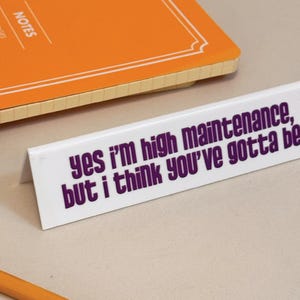 May include: A white desk sign with the text "yes I'm high maintenance, but I think you've gotta be" in purple. An orange notebook and a pencil are also in the image.