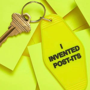 Pu&ograve; includere: Un portachiavi giallo a forma di chiave di motel con il testo "Ho inventato i Post-it" in lettere nere. Il portachiavi &egrave; attaccato a una chiave dorata.