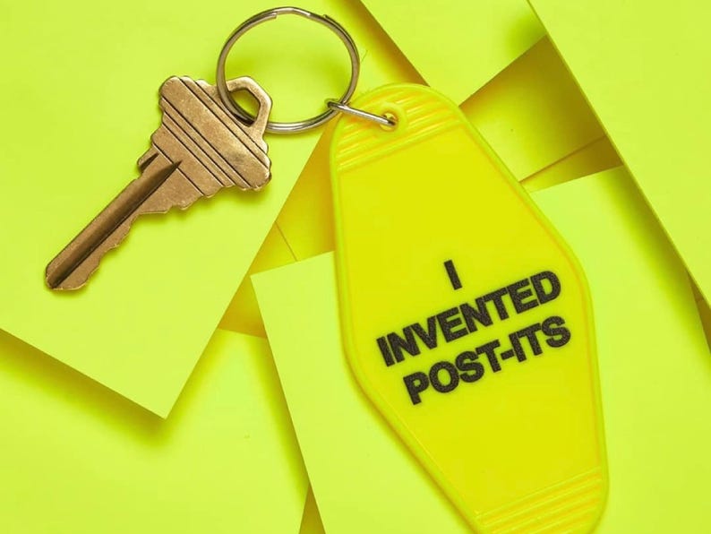 Pu&ograve; includere: Un portachiavi giallo a forma di chiave di motel con il testo "Ho inventato i Post-it" in lettere nere. Il portachiavi &egrave; attaccato a una chiave dorata.