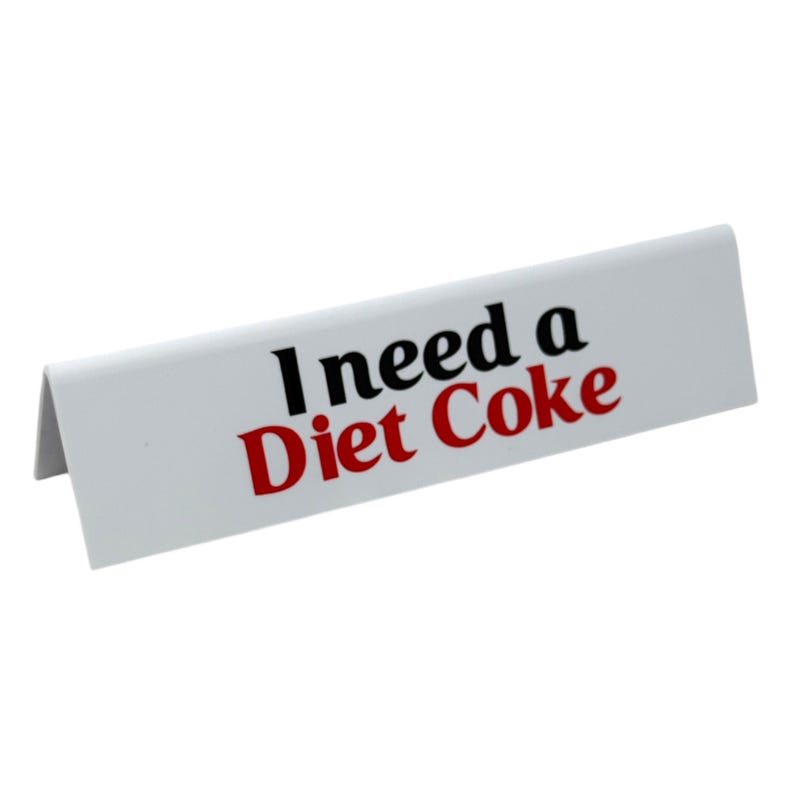 May include: A white, angled sign with the text "I need a Diet Coke" in black and red. The sign is a simple, rectangular shape, designed to stand upright.