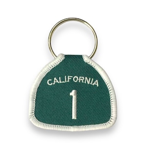 May include: Green and white California Route 1 keychain with a silver key ring. The keychain is shaped like a highway sign with the number 1 in the center.